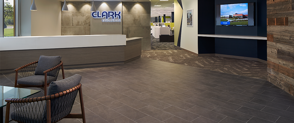 lobby with dark stone tile floors and walls, large desk of wood and white panel, Clark Engineering neon sign, TV, chairs and table, view into desk space